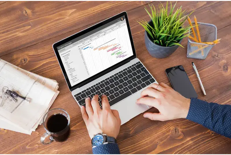 A desk with a laptop showing a Gantt chart, surrounded by a planner, coffee, smartphone, pen, and a potted plant.